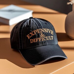 Peut inclure: Une casquette de baseball noire avec une visière incurvée et un texte brodé de couleur beige indiquant EXPENSIVE & DIFFICULT. La casquette présente une construction à six panneaux avec des œillets d'aération et des coutures visibles.