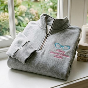 Könnte beinhalten: Ein graumeliertes Sweatshirt mit kurzem Reißverschluss, das eine Stickerei einer hellblauen Katzenaugenbrille über dem rosa gestickten Text zeigt: Abbey, Licensed Optician, ABOC. Das Kleidungsstück hat einen hohen Kragen mit einem Metallreißverschluss.