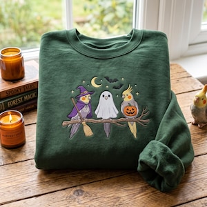 May include: A dark green crewneck sweatshirt featuring an embroidered design of two cockatiel birds and a ghost perched on a branch. One bird wears a purple witch hat and cape with a broom, while the other holds a jack-o'-lantern pumpkin.