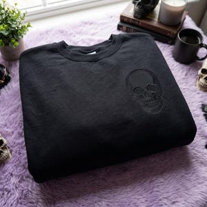 Puede incluir: Una sudadera negra de cuello redondo con un diseño de calavera bordado en negro tonal en la parte superior izquierda del pecho. La prenda tiene cuello, puños y dobladillo acanalados, con un bordado que muestra detalles precisos.