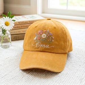 以下が含まれることがあります： ピンク、白、紫の色合いで野草の花束が刺繍されたマスタードイエローの野球帽。花のデザインの下には、白い筆記体でOmaという文字がフロントパネルに刺繍されています。