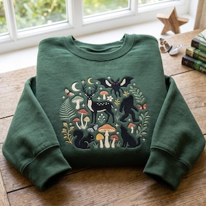 Puede incluir: Una sudadera de cuello redondo verde oscuro con un diseño bordado central de un ciervo negro, una ardilla, un lobo, una polilla y un sasquatch rodeados de varios hongos coloridos, helechos, mariposas, lunas crecientes y estrellas pequeñas.