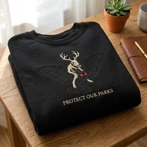 May include: A black crewneck sweatshirt featuring an embroidered graphic of a skeletal deer-like creature riding a dark winged moth with bright red eyes. The text PROTECT OUR PARKS is embroidered in a light beige colour below the central graphic.