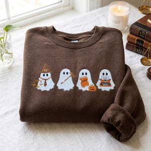 Puede incluir: Una sudadera de cuello redondo color marrón oscuro con un diseño bordado de cuatro fantasmas blancos. El primer fantasma lleva sombrero, corbata y sostiene una balanza. El segundo sostiene un mazo. El tercero sostiene un libro con la palabra LAW junto a una calabaza. El cuarto sostiene una máquina de escribir.