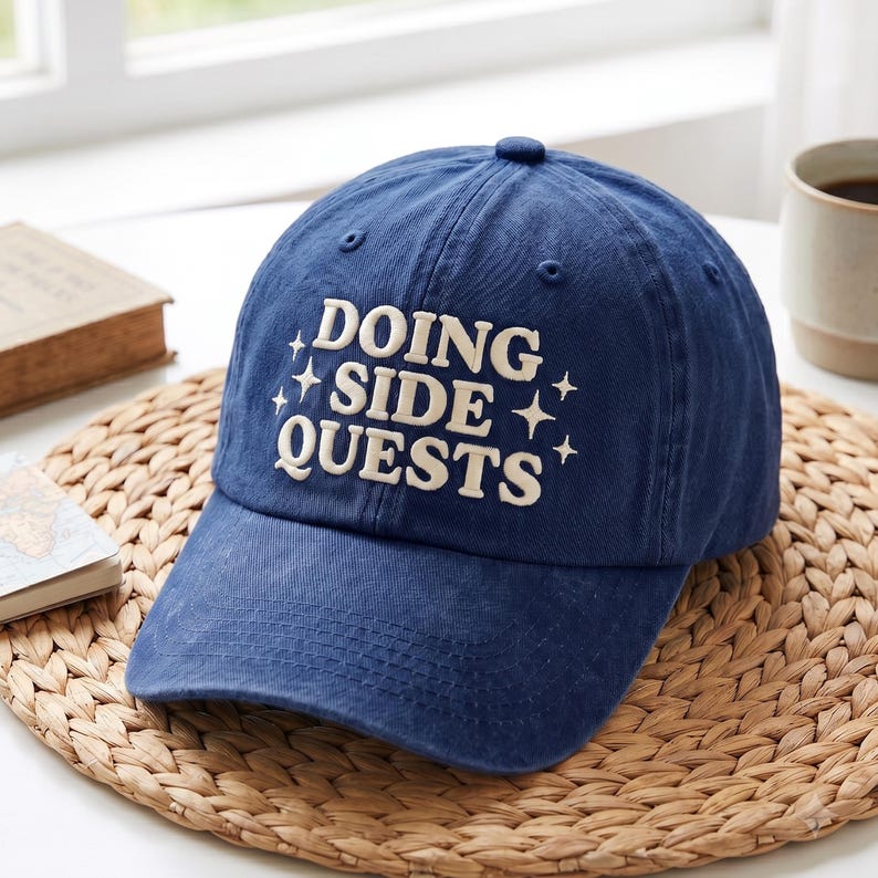 May include: A navy blue baseball cap featuring the embroidered white text DOING SIDE QUESTS with small four-pointed star icons on the front panel. The cap has a structured crown, a curved brim, and stitched eyelets for ventilation.