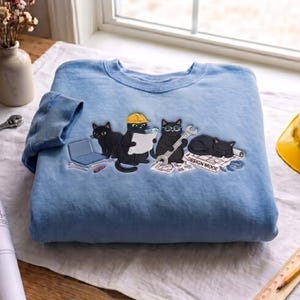Op de afbeelding: Een lichtblauw sweatshirt met ronde hals en een geborduurde afbeelding van vier zwarte katten. Eén kat draagt een gele veiligheidshelm en houdt een bouwtekening vast, een andere draagt een bril en houdt een moersleutel vast, met de tekst DESIGN MODE.