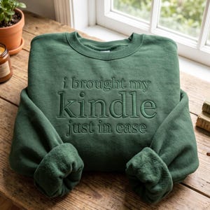 以下が含まれることがあります： フォレストグリーンのクルーネックトレーナー。同系色の緑色の糸で「i brought my kindle just in case」という文字が刺繍されています。長袖で袖口と襟元はリブ編みになっており、シンプルで無地のデザインが特徴です。
