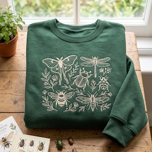 Può includere: Una felpa girocollo verde bosco scuro con un ricamo color crema raffigurante vari insetti, tra cui una falena, una libellula, uno scarabeo, una coccinella e un'ape, intervallati da delicati rametti botanici ed elementi floreali.