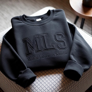 Puede incluir: Una sudadera negra de cuello redondo con las letras MLS bordadas en una fuente universitaria grande, con el texto Medical Laboratory Scientist bordado en una fuente más pequeña justo debajo de las letras grandes en el pecho.