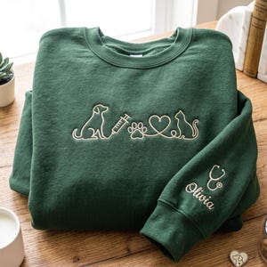 May include: A dark green crewneck sweatshirt featuring an embroidered line art design on the chest of a dog, a syringe, a paw print, a heart, and a cat. The sleeve is embroidered with a stethoscope icon and the name Olivia in cursive script.