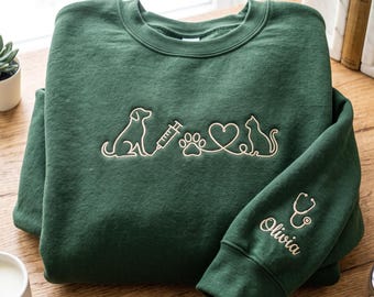 May include: A dark green crewneck sweatshirt featuring an embroidered line art design on the chest of a dog, a syringe, a paw print, a heart, and a cat. The sleeve is embroidered with a stethoscope icon and the name Olivia in cursive script.