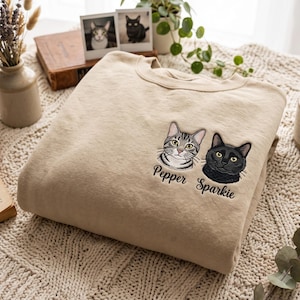 Puede incluir: Una sudadera beige de cuello redondo con retratos bordados de dos gatos en el pecho. Un gato es atigrado gris con el nombre Pepper bordado debajo, y el otro es un gato negro sólido con el nombre Sparkie bordado debajo.