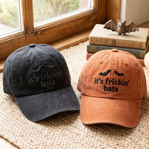 Peut inclure: Deux casquettes de baseball, une gris anthracite et une orange brûlé, arborant chacune le texte brodé en noir 'it's frickin' bats' sous deux petites silhouettes de chauves-souris. Les casquettes ont une calotte structurée et une visière.
