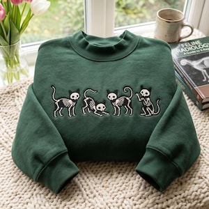 Op de afbeelding: Een donkergroene sweater met ronde hals en een geborduurd ontwerp van vier zwarte katten met witte skeletpatronen. De katten zijn afgebeeld in verschillende houdingen, waaronder staand, uitrekkend en zittend, gecentreerd op de borst.