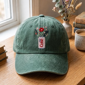 May include: A forest green baseball cap featuring an embroidered design on the front. The embroidery depicts a light pink soda can labeled Diet Coke, which serves as a vase for a small bouquet of colorful flowers including a prominent red bloom.