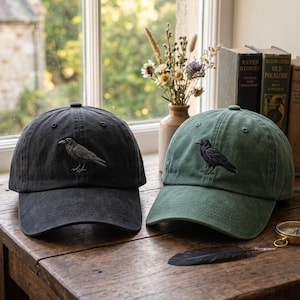 May include: Two baseball caps, one in charcoal gray and one in sage green, each featuring an embroidered black crow on the front panel. The caps have a structured crown, a curved brim, and a fabric strap with a metal buckle at the back.