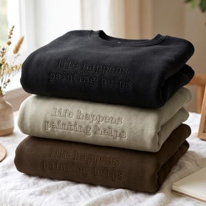 Könnte beinhalten: Ein Stapel aus drei Rundhals-Sweatshirts in Schwarz, Beige und Dunkelbraun. Jedes Sweatshirt ist mit dem gestickten Text "life happens painting helps" in passender Garnfarbe mittig auf der Brustpartie versehen.