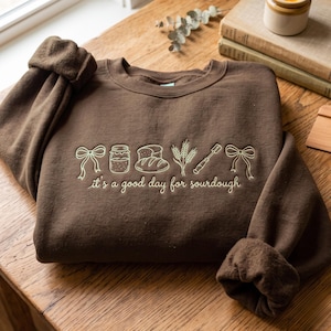 May include: A dark brown crewneck sweatshirt featuring light-colored embroidery of two bows, a jar, a loaf of bread, wheat stalks, and a bread lame. The text "it's a good day for sourdough" is embroidered in a cursive font below the icons.