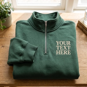 Puede incluir: Una sudadera verde oscuro con cremallera de un cuarto, cuello alto y cremallera de metal plateado. En el pecho aparece el texto bordado YOUR TEXT HERE en color beige claro. La prenda tiene mangas largas con puños de canalé.
