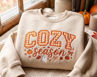 May include: A beige crewneck sweatshirt featuring embroidered text that reads COZY season. The word COZY is in orange and white gingham block letters, while season is in orange cursive script, surrounded by small pumpkin, ghost, and leaf icons.