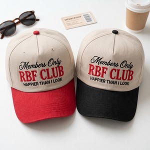 Könnte beinhalten: Zwei Baseballkappen mit beigen Kronen und der Stickerei Members Only RBF CLUB HAPPIER THAN I LOOK. Eine Kappe hat einen roten Schirm und Knopf, die andere einen schwarzen Schirm und Knopf. Beide Kappen bestehen aus fünf Paneelen.