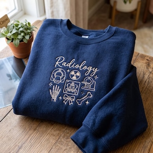 May include: A navy blue crewneck sweatshirt featuring white embroidered line art graphics and the text "Radiology". The design includes icons of a CT scanner, a radiation symbol, an X-ray of a pelvis, a hand skeleton, a bow, and a bone.