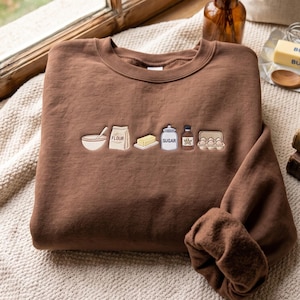 Könnte beinhalten: Ein braunes Sweatshirt mit Rundhalsausschnitt und einer Stickerei von Backzutaten auf der Brust. Das Motiv zeigt eine Rührschüssel, eine Tüte mit der Aufschrift FLOUR, ein Stück Butter, ein Glas mit der Aufschrift SUGAR, eine Flasche und Eier.