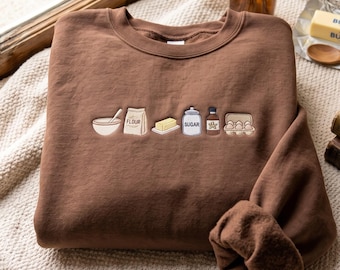 May include: A brown crewneck sweatshirt featuring an embroidered design of baking ingredients across the chest. The embroidery includes a mixing bowl, a bag labeled FLOUR, a stick of butter, a jar labeled SUGAR, a bottle, and a carton of eggs.