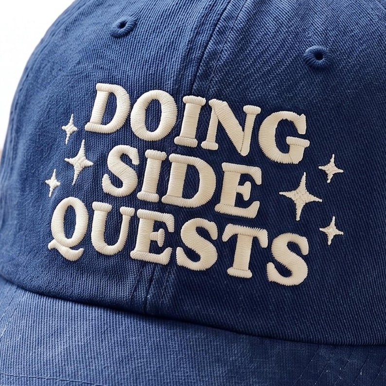 May include: A navy blue baseball cap featuring the embroidered text DOING SIDE QUESTS in a cream-colored font. The design is accented with four-pointed star shapes on both sides of the text, all stitched in a matching cream thread.