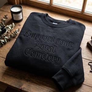 May include: A black crewneck sweatshirt featuring the text Support your Local Cemetery embroidered in a matching black thread using a gothic-style font. The garment has long sleeves and a ribbed collar, cuffs, and hem, showcasing a monochromatic design.