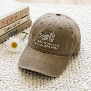Puede incluir: Una gorra de béisbol marrón con un gráfico bordado en blanco de una pila de libros y un café helado, junto al texto "i'll take my books hot and my coffee iced." La gorra tiene una corona estructurada, una visera curva y un botón superior.
