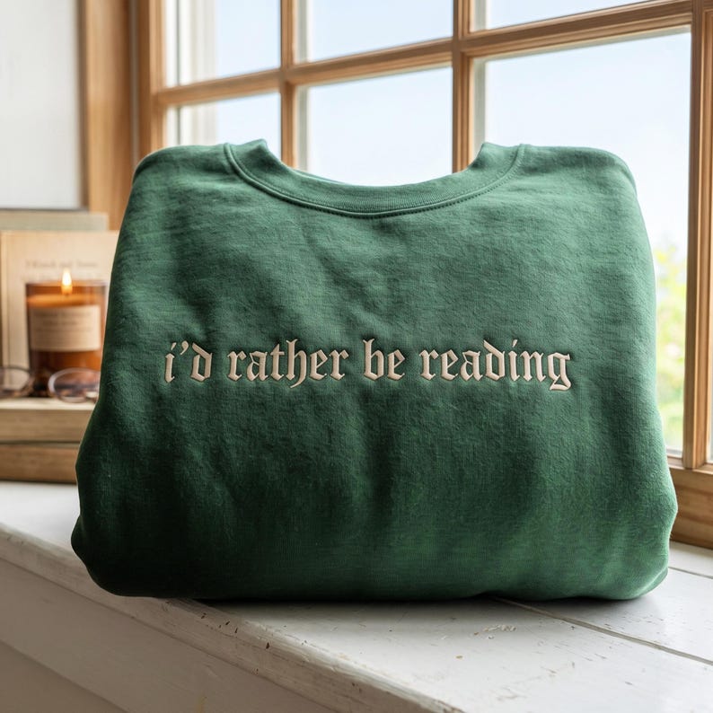 May include: A forest green crewneck sweatshirt featuring the embroidered text "i'd rather be reading" in a cream-colored gothic-style font centered on the chest. The garment has long sleeves, a ribbed neckline, and a soft, solid-colored fabric.
