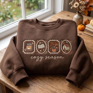 Peut inclure: Un sweat-shirt marron foncé à col rond avec des broderies représentant une bougie, un livre ouvert, un vase avec des feuilles d'automne et une petite maison, encadrés par des bordures décoratives. Le texte "cozy season" est brodé en blanc.