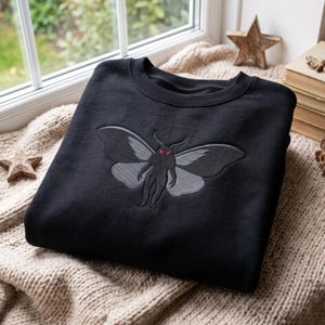 Op de afbeelding: Een zwart sweatshirt met ronde hals en een grote geborduurde afbeelding op de borst van een mot-achtig wezen met een mensachtig lichaam, donkergrijze vleugels en felrode ogen. Het kledingstuk heeft een geribbelde kraag.