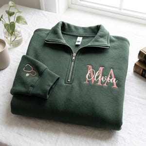 Puede incluir: Una sudadera verde oscuro con cremallera de un cuarto, cremallera plateada, un estetoscopio bordado en oro rosa en el puño de la manga y el nombre Olivia bordado en cursiva sobre letras grandes en bloque color oro rosa MA.
