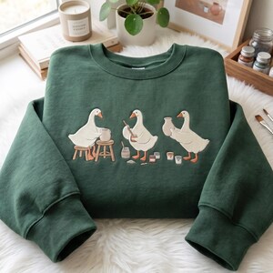 以下が含まれることがあります： 陶芸をする3羽の白いガチョウが刺繍されたダークグリーンのクルーネックトレーナー。ガチョウはスツールに座り、道具を使い、粘土の壺を持っており、その周りには小さな瓶やボウルが配置されている様子が描かれている。