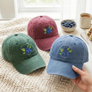 May include: Three baseball caps in green, maroon, and blue, each featuring an embroidered design of three blueberries with green leaves on the front panel. The caps have a washed, textured fabric finish and a structured six-panel construction.