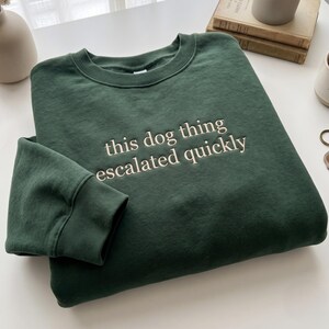 May include: A dark forest green crewneck sweatshirt featuring the embroidered text "this dog thing escalated quickly" in a light cream color. The garment has long sleeves with ribbed cuffs and a ribbed neckline, showcasing a clean, simple design.