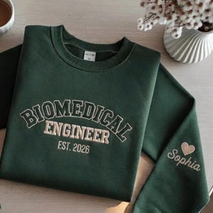 Peut inclure: Un sweat-shirt à col rond vert foncé avec le texte brodé BIOMEDICAL ENGINEER EST. 2026 dans une police universitaire crème. La manche présente le prénom Sophia brodé en crème accompagné d'une petite icône de cœur pailleté.