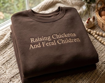 Può includere: Una felpa girocollo marrone scuro con il testo ricamato "Raising Chickens And Feral Children" in filo color marrone chiaro. Il capo presenta maniche lunghe, un colletto a coste e una struttura in tessuto morbido a tinta unita.