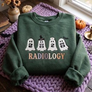 May include: A dark green crewneck sweatshirt featuring an embroidered graphic of four white ghosts holding medical X-rays. One ghost wears a witch hat, another a nurse cap. The word RADIOLOGY is embroidered below in multicolored pastel letters.