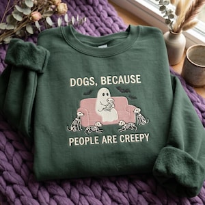 Op de afbeelding: Een donkergroene sweater met ronde hals en een geborduurde afbeelding van een witte geest op een roze bank, die een skelet-hond vasthoudt, omringd door vier skelet-honden en vleermuizen, met de tekst DOGS, BECAUSE PEOPLE ARE CREEPY.