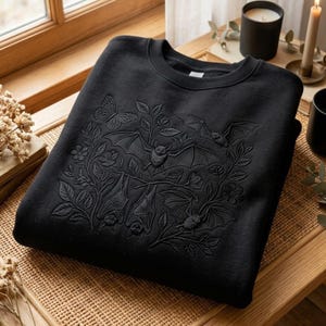 Peut inclure: Un sweat-shirt noir à col rond orné d'une broderie ton sur ton représentant une scène botanique. Le motif comprend plusieurs chauves-souris en vol et suspendues, entremêlées de branches fleuries détaillées et d'un papillon.