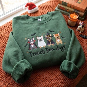Könnte beinhalten: Ein dunkelgrüner Rundhals-Pullover mit einer Stickerei von vier französischen Bulldoggen in Halloween-Kostümen, darunter ein Vampir, ein Geist, eine Hexe und ein Kürbis, sowie dem Schriftzug "French Boo-dogs" und kleinen schwarzen Fledermäusen.