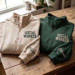 Puede incluir: Una sudadera beige con cremallera de cuarto, texto bordado en verde oscuro SOCIAL WORKER y el nombre Olivia en la manga, junto a una sudadera verde oscuro con texto bordado en beige SOCIAL WORKER y el nombre Harper en la manga.