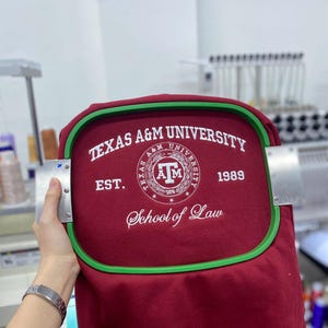 May include: A maroon fabric garment secured in a green rectangular embroidery hoop. The fabric features white embroidered text reading TEXAS A&M UNIVERSITY, EST. 1989, and School of Law, surrounding a circular Texas A&M University logo emblem.