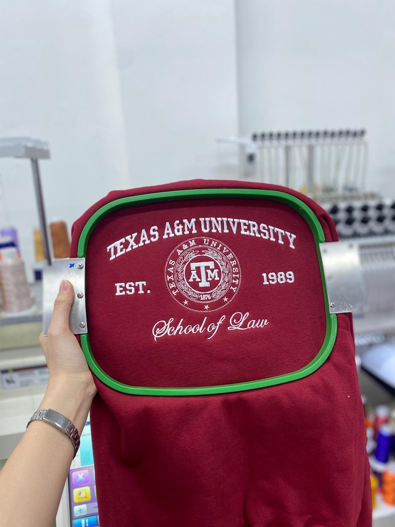 May include: A maroon fabric garment secured in a green rectangular embroidery hoop. The fabric features white embroidered text reading TEXAS A&M UNIVERSITY, EST. 1989, and School of Law, surrounding a circular Texas A&M University logo emblem.