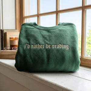 May include: A forest green crewneck sweatshirt featuring the embroidered text "i'd rather be reading" in a cream-colored gothic font centered on the chest. The garment has long sleeves and a ribbed neckline, showcasing a simple and clean design.