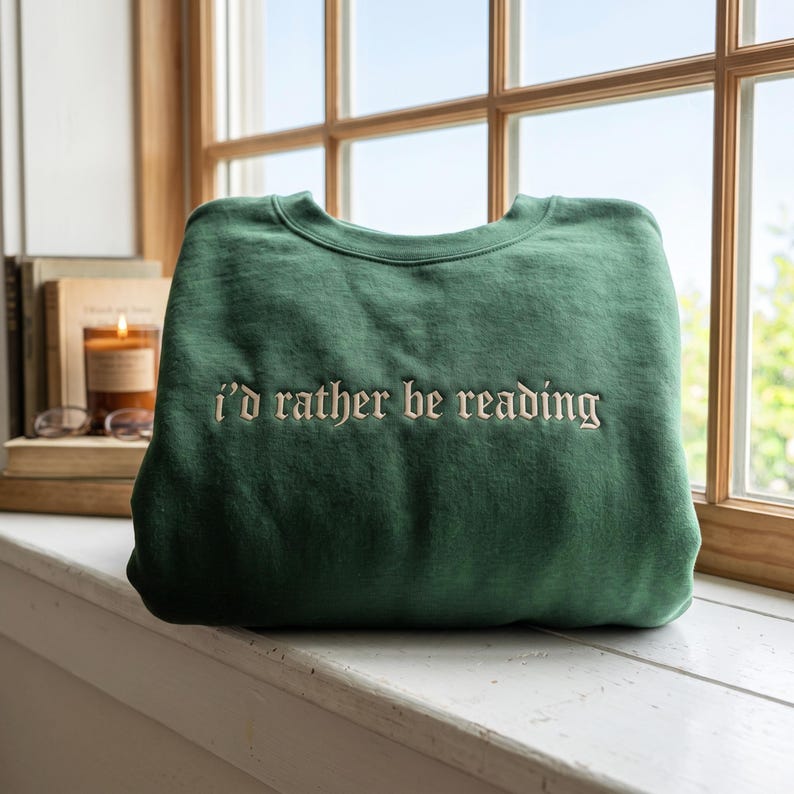 May include: A forest green crewneck sweatshirt featuring the embroidered text "i'd rather be reading" in a cream-colored gothic font centered on the chest. The garment has long sleeves and a ribbed neckline, showcasing a simple and clean design.