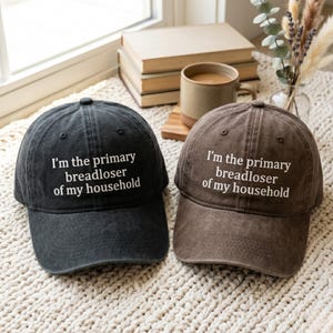 May include: Two baseball caps, one in charcoal black and one in brown, both featuring a distressed fabric finish. Each cap is embroidered with the white text "I'm the primary breadloser of my household" centered on the front panel above the brim.